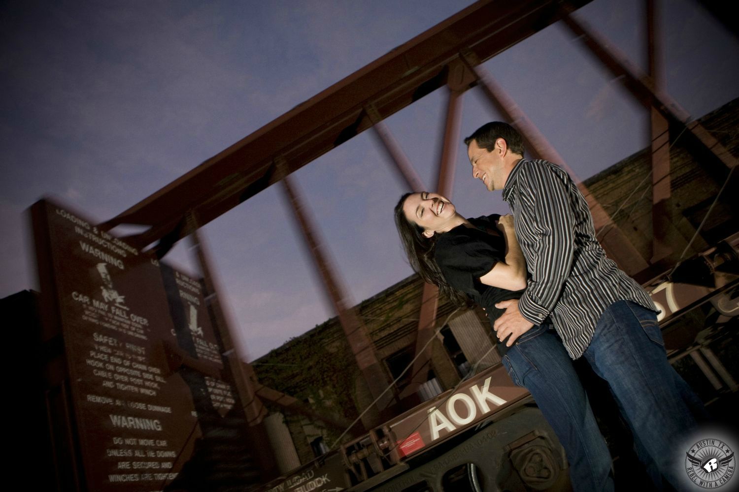 Dark haired laughing girl wearing a black button up short sleeve blouse with blue jeans is held by a dark haired guy wearing a black and white striped dress shirt and blue jeans laughs as a freight train passes by in the background at dusk with a dark blue cloudy night sky in this fun engagement portrait near the Amtrak Station in Austin, Texas. 